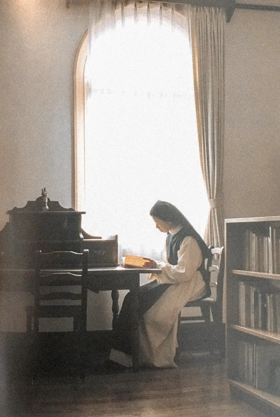A nun writing at a desk beside a bright window.